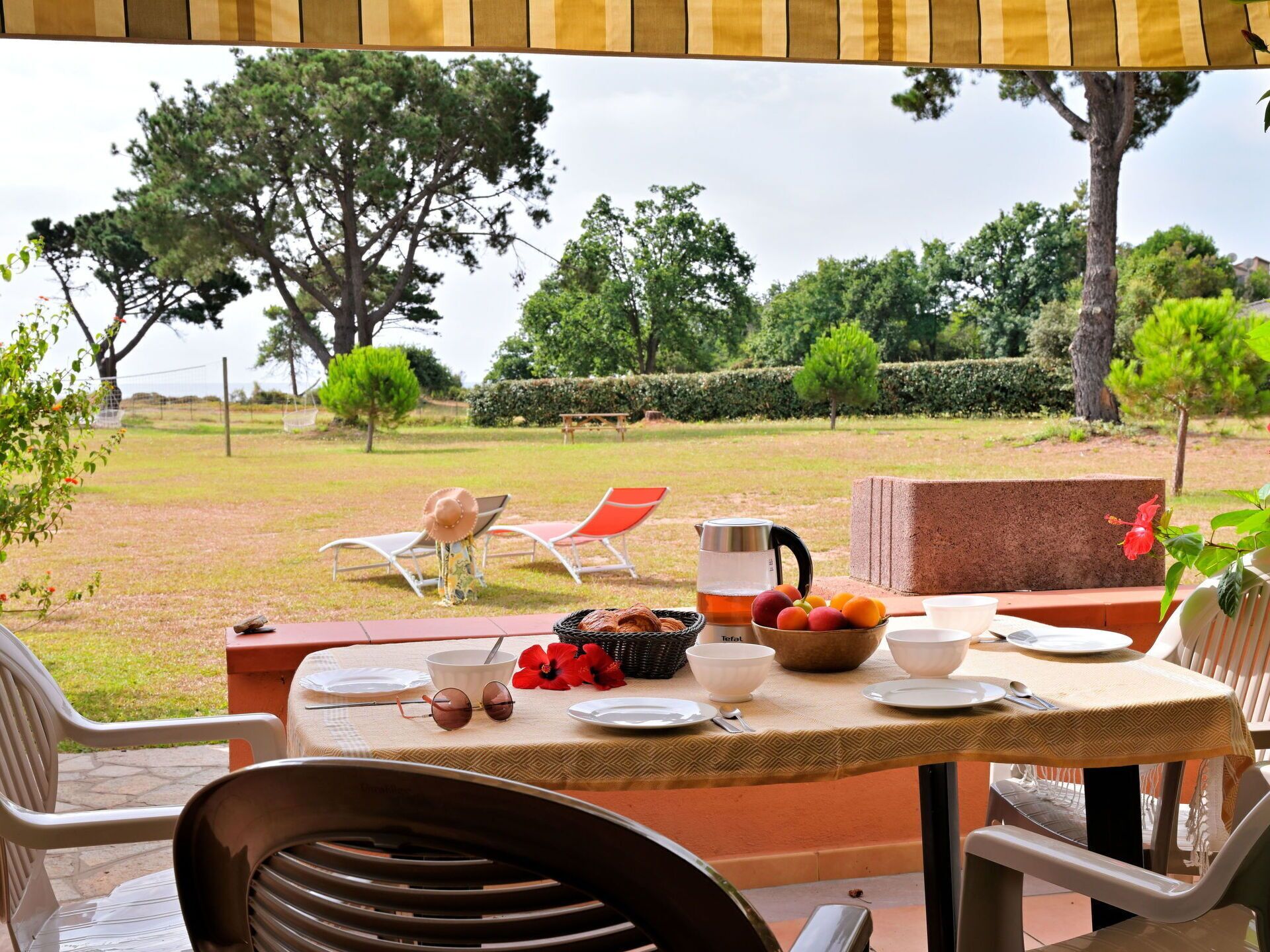 An outdoor dining area with a table set for two, surrounded by chairs, with a view of a grassy garden and trees.