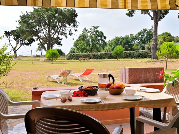 An outdoor dining area with a table set for two, surrounded by chairs, with a view of a grassy garden and trees.