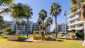 A playground with a yellow slide and climbing structure, surrounded by palm trees and modern apartment buildings. - Cosy apartment for 4 people with A/C, pool, WIFI, TV, terrace and pets allowed (Torredembarra)