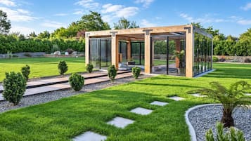 Una estructura moderna al aire libre con paredes de vidrio, rodeada por un césped y jardín cuidados.