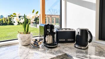 Uma bancada de cozinha com cafeteira, torradeira e xícara, um vaso com flores e vista para uma paisagem verde pela janela.