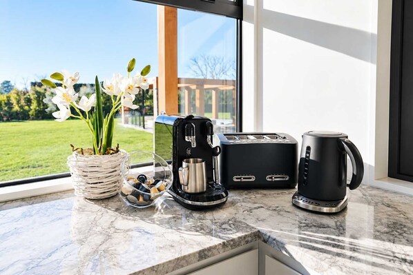 A kitchen counter with a coffee maker, toaster, and mug, a vase with flowers, and a view of a green landscape through a window.