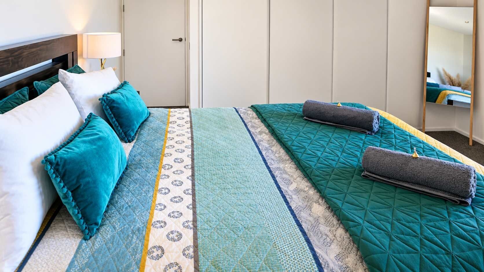 Una habitación de hotel con cama, almohadas, mesita de noche con lámpara y un espejo.