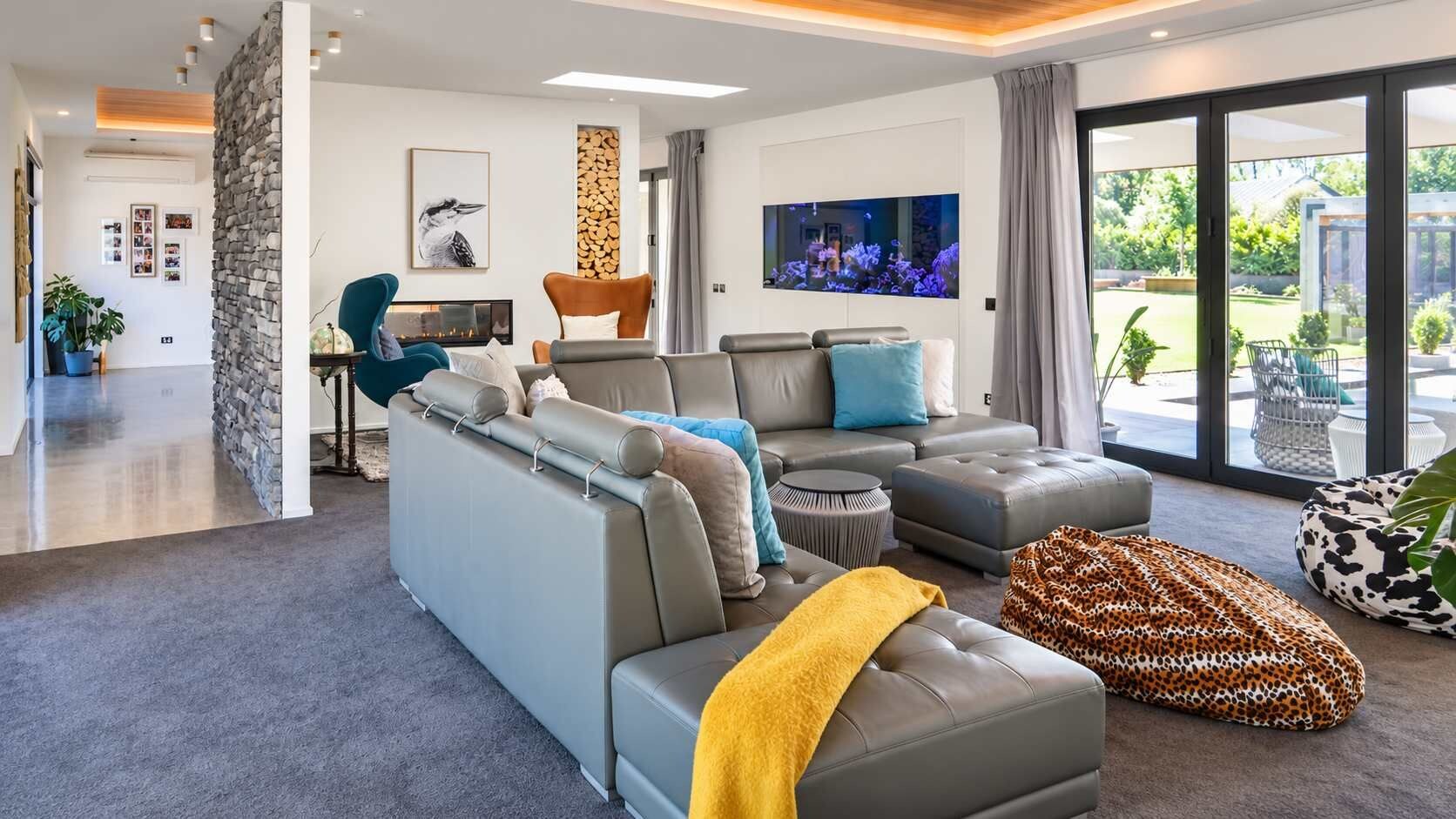 A modern living room with a grey sectional sofa, a coffee table, and a leopard print pouf. There is a flat-screen TV mounted on the wall, and a stone wall feature is visible.