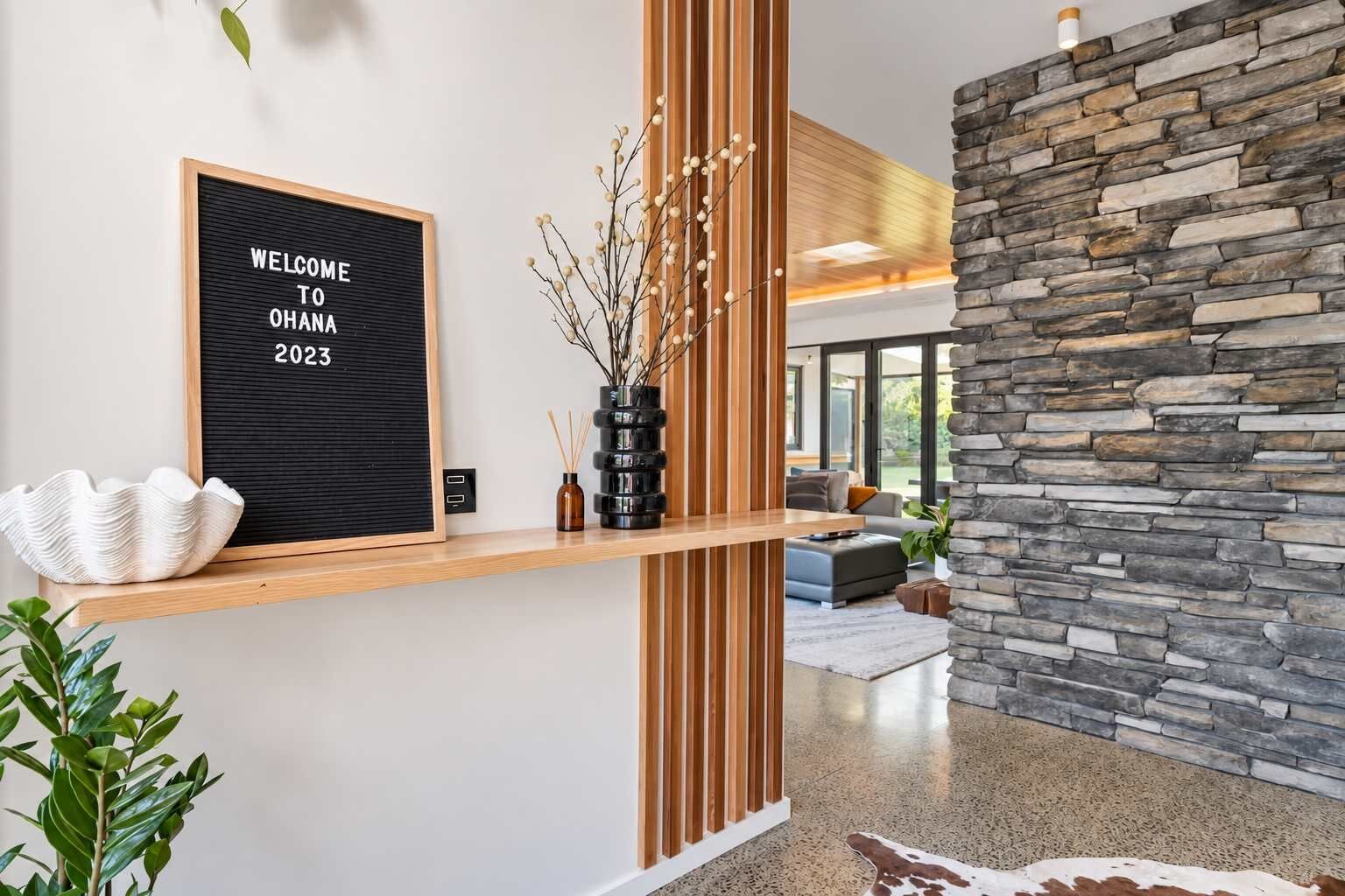 Una moderna area reception con un cartello di benvenuto, uno scaffale in legno con oggetti decorativi, un muro in pietra e uno scorcio di un soggiorno con un divano e una pianta.