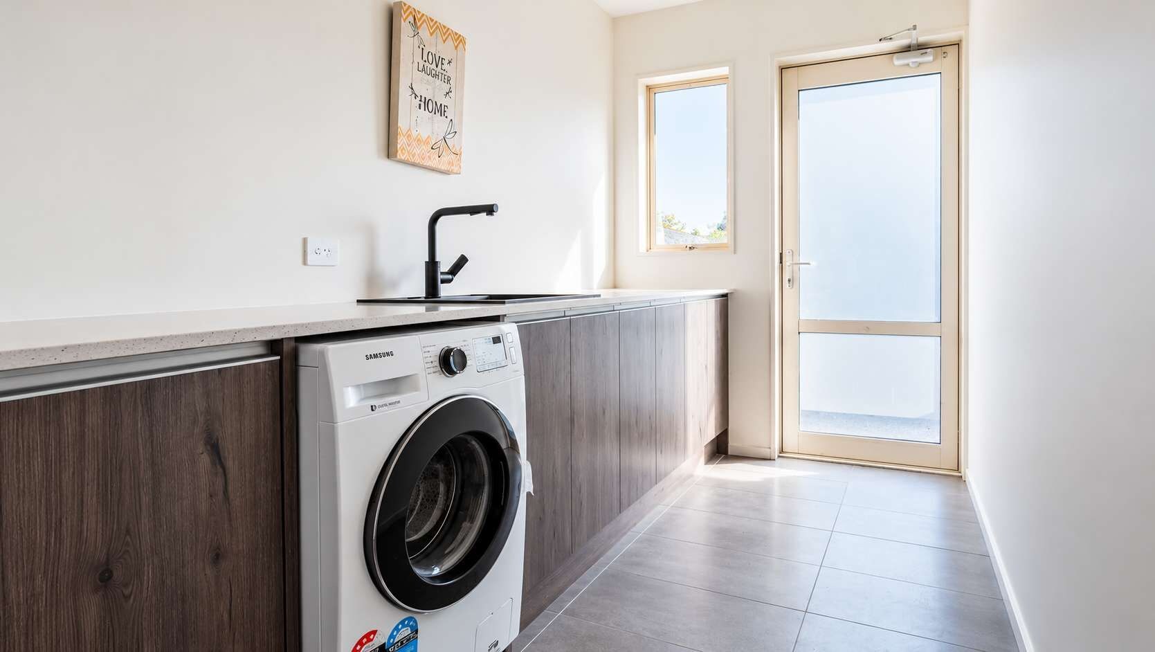 A laundry room with a Samsung washing machine, a wooden cabinet, a wall-mounted towel rack, and a door leading to a balcony.