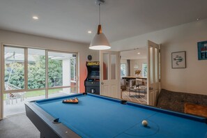 A pool table with a blue felt surface, a hanging pendant light, and a view of the outdoors through large windows. - Hidden Gem of Christchurch - Sleeps 12 (Christchurch)