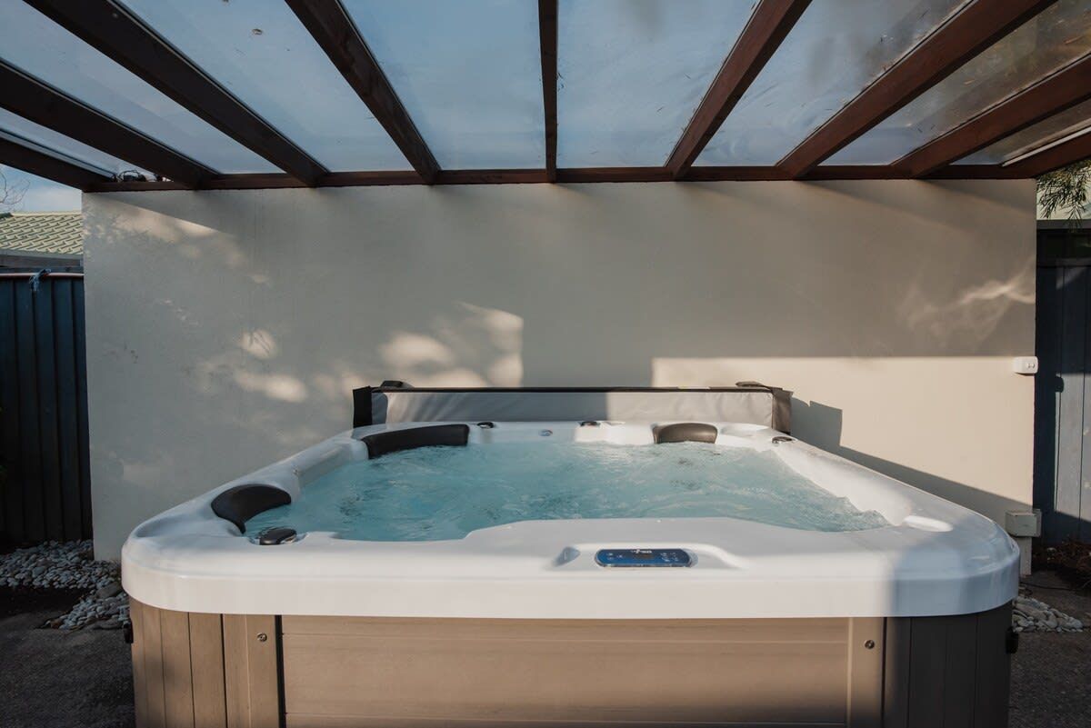 Un jacuzzi bajo una pérgola con estructura de madera y una pared blanca al fondo.