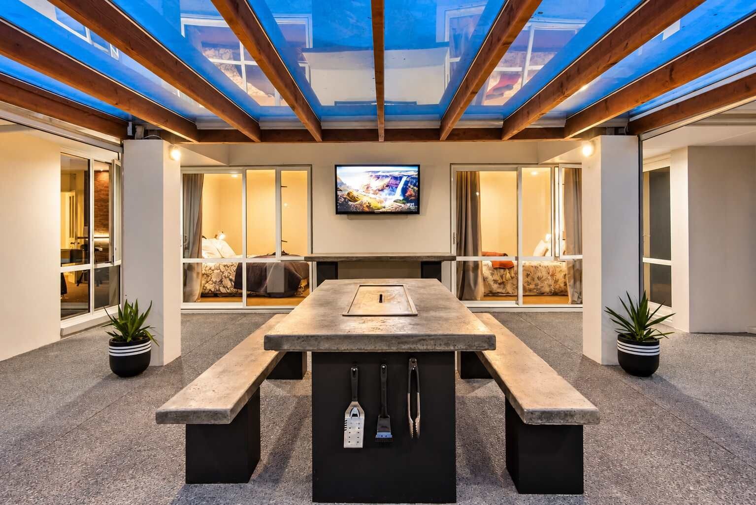 A modern outdoor dining area with a stone table and benches, a television mounted on the wall, and potted plants.