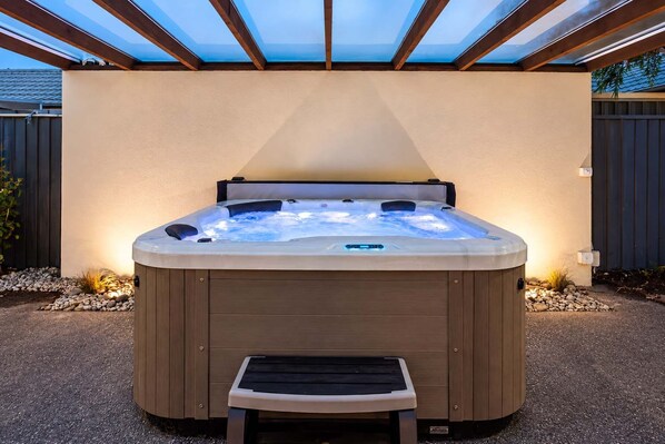 A hot tub with a wooden bench in front, surrounded by a patio area with a pergola overhead.