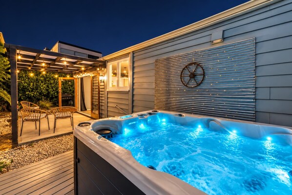 Un jacuzzi con cubierta de madera y sillas, una pérgola con luces colgantes y un muro con listones horizontales y una decoración de volante.