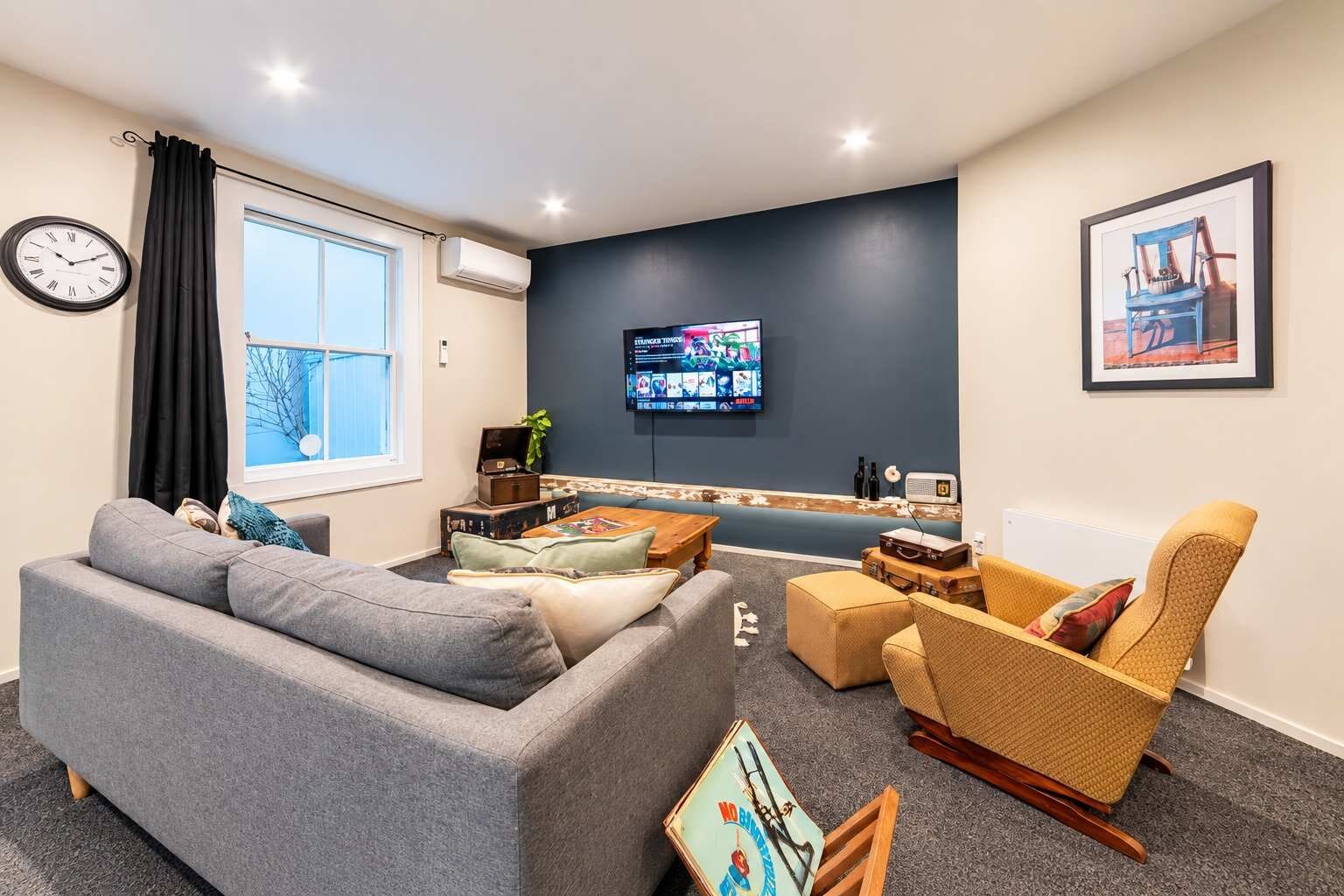 A living room with a grey sofa, a yellow armchair, a wooden coffee table, and a flat-screen TV mounted on the wall.