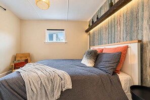 Een slaapkamer met een bed, een stoel, een raam en een wandplank met flessen.