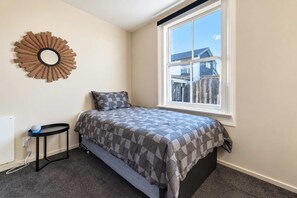 A bedroom with a bed, a round mirror, a small table, and a window with a view of a house. - Richmond Cottage | 3BR + Hot Tub | Parking (Christchurch)