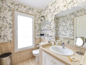 Casa de banho com papel de parede floral, sanita, lavatório com espelho e prateleiras com artigos de higiene pessoal.