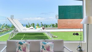 A modern living room with a sofa, two decorative pillows, a small table with drinks, and a view of a tennis court and greenery. - Wonderful private villa for 6 people with private pool and TV (Arucas)
