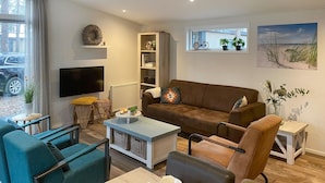 A modern living room with a brown sofa, teal armchair, and a coffee table. A TV is mounted on the wall, and there are plants and artwork on the shelves.