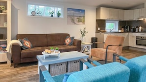 A modern living room with a brown sofa, a blue armchair, and a small table with candles and plants. There is a kitchen area with white cabinets and a dining table.