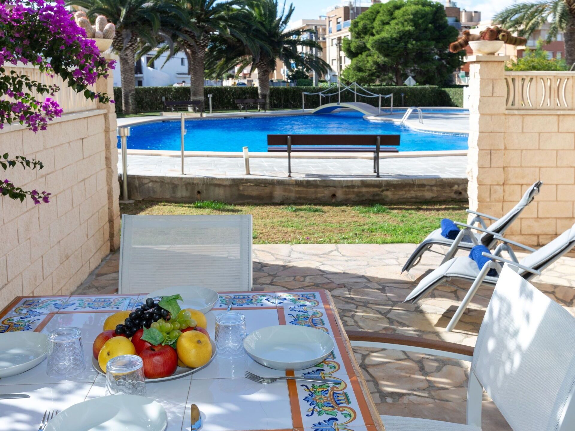 Una mesa de comedor con fruteros, platos y cubiertos, con vista a una piscina y palmeras.