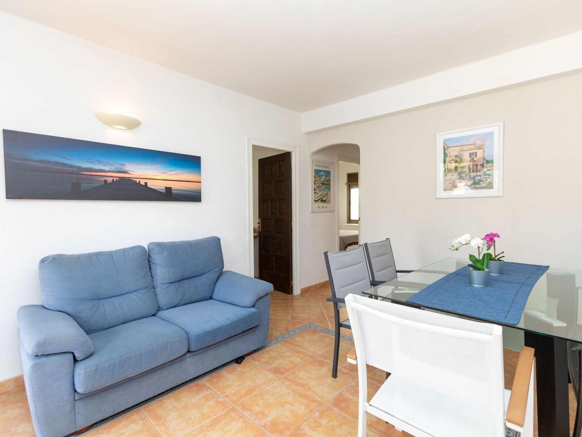 Un espace de vie comprenant un canapé bleu, une table à manger en verre avec des chaises, et un tableau représentant un coucher de soleil accroché au mur.