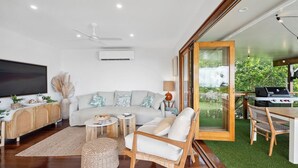A modern living room with a grey sofa, a wooden coffee table, and a TV. There's a dining area with a table and chairs, and a large window overlooking greenery. - TreasureView Beach - Three bedroom option #9a (Coolum Beach)