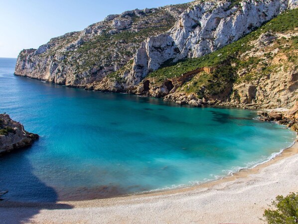 Experience the serene coastal landscape with turquoise sea and sandy beach, surrounded by natural rocky cliffs and sparse vegetation.