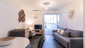 A modern living room with a grey sofa, a dining table, and a TV. There is a balcony with a railing and a view of the sky. - Lovely apartment for 2 guests with WIFI, A/C, pool, TV and terrace (Porto-vecchio)