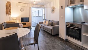 A modern kitchen with a built-in oven, a dining area with a round table and chairs, a grey sofa, and a large window offering a view of the outdoors. - Lovely apartment for 2 guests with WIFI, A/C, pool, TV and terrace (Porto-vecchio)