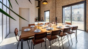 A spacious dining area with a long wooden table set for six, surrounded by leather chairs. The room features exposed brick walls, large windows offering a view of the countryside, and hanging pendant lights. - Beautiful private villa for 14 guests with private pool, WIFI, hot tub, A/C, TV and patio (Poirino)
