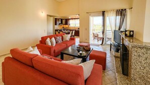 Una sala de estar con sofás rojos, una mesa de centro y un televisor. Se observa una cocina al fondo, con armarios y una ventana.