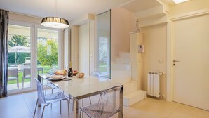 A modern kitchen with a large island, clear chairs, and a view of the garden through glass doors.