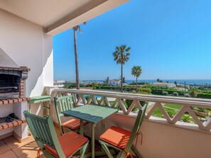 Un balcón con una parrilla, sillas y una mesa, con vistas a la costa, palmeras y casas.