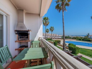 A balcony with a grill, a table, and chairs, overlooking a swimming pool and palm trees.