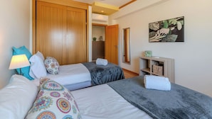 A bedroom with two beds, a wooden wardrobe, a bedside table with a lamp, a mirror, and a wall-mounted artwork.