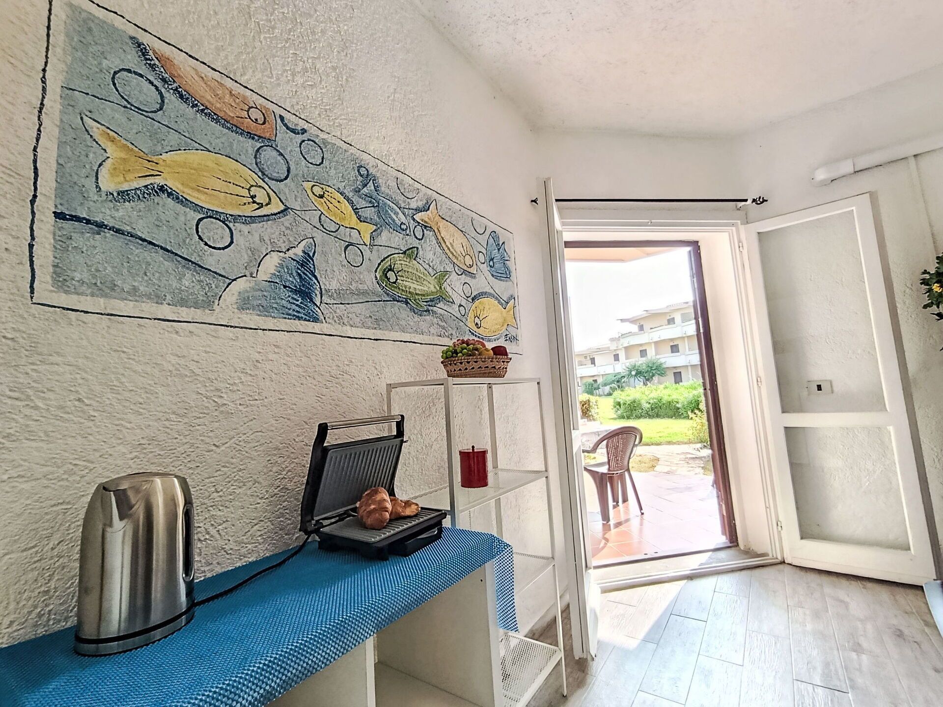 Une pièce avec un coin cuisine, un tableau représentant des poissons, un panier de fruits et une vue sur l’extérieur.