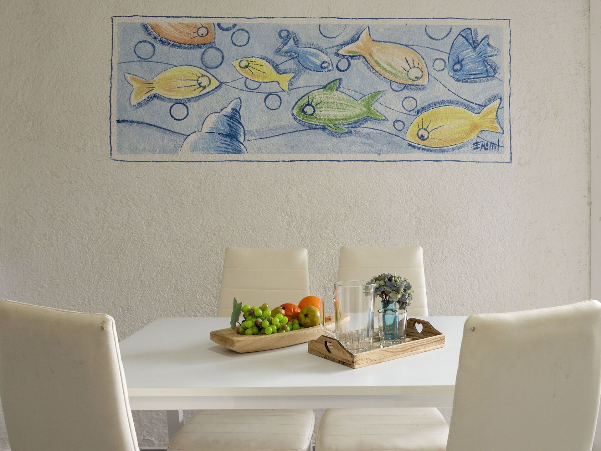 A dining area with a white table, two chairs, a wooden tray with fruits and glasses, and a wall painting of fish.