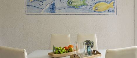 A dining area with a white table, two chairs, a wooden tray with fruits and glasses, and a wall painting of fish.