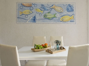 A dining area with a white table, two chairs, a wooden tray with fruits and glasses, and a wall painting of fish. - Cosy apartment for 4 people with WIFI, A/C, pool, TV, terrace and pets allowed (Golfo Aranci)