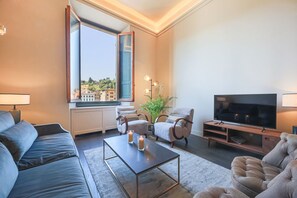 A modern living room with a sofa, armchairs, a coffee table, a TV, and a large window with a view of buildings. - Apartments Florence - Palazzo Benci with river view · Palazzo Benci with river v (Firenze)