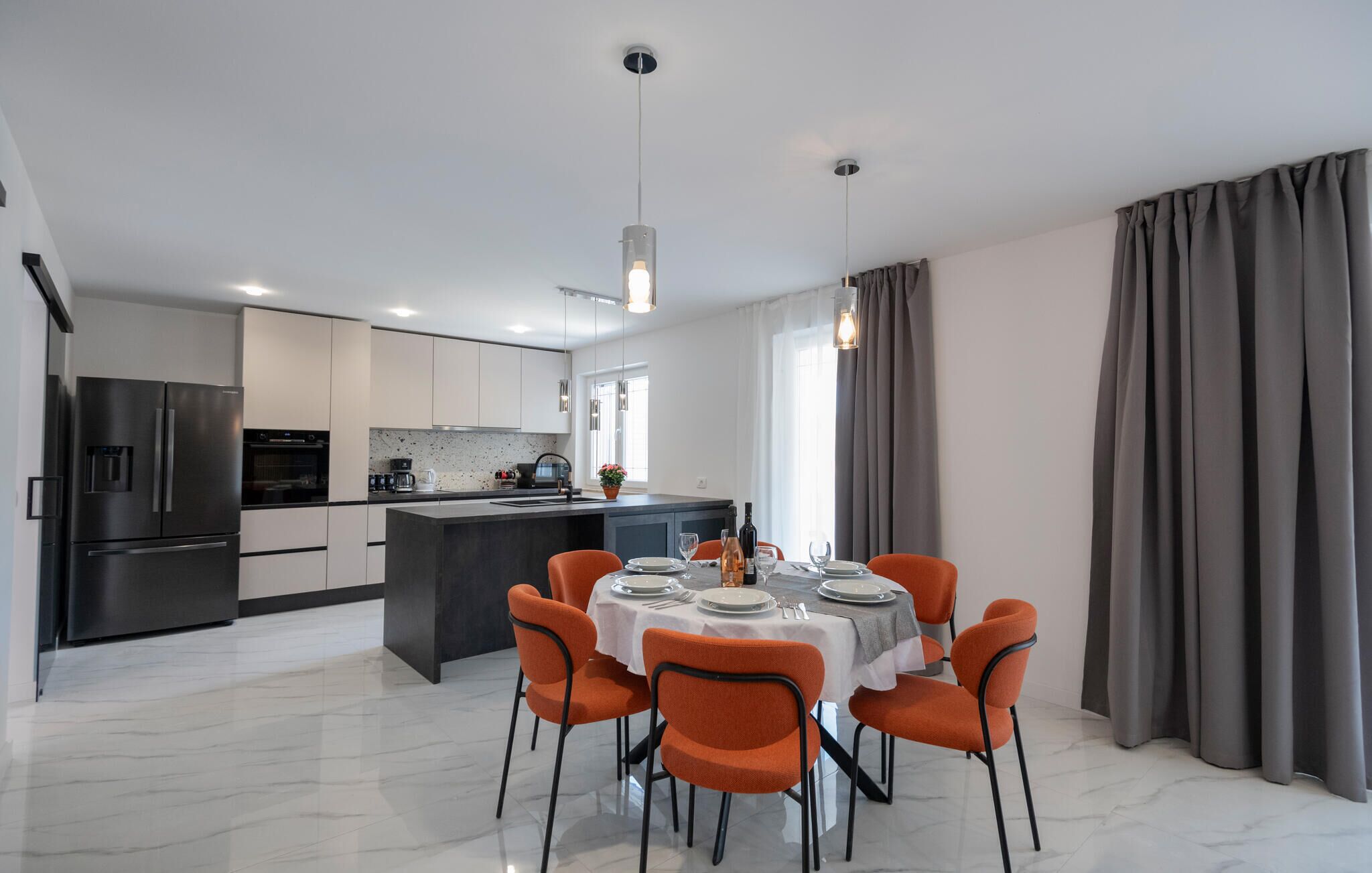 A modern kitchen with a central island, a dining area with orange chairs, and a large window with curtains.