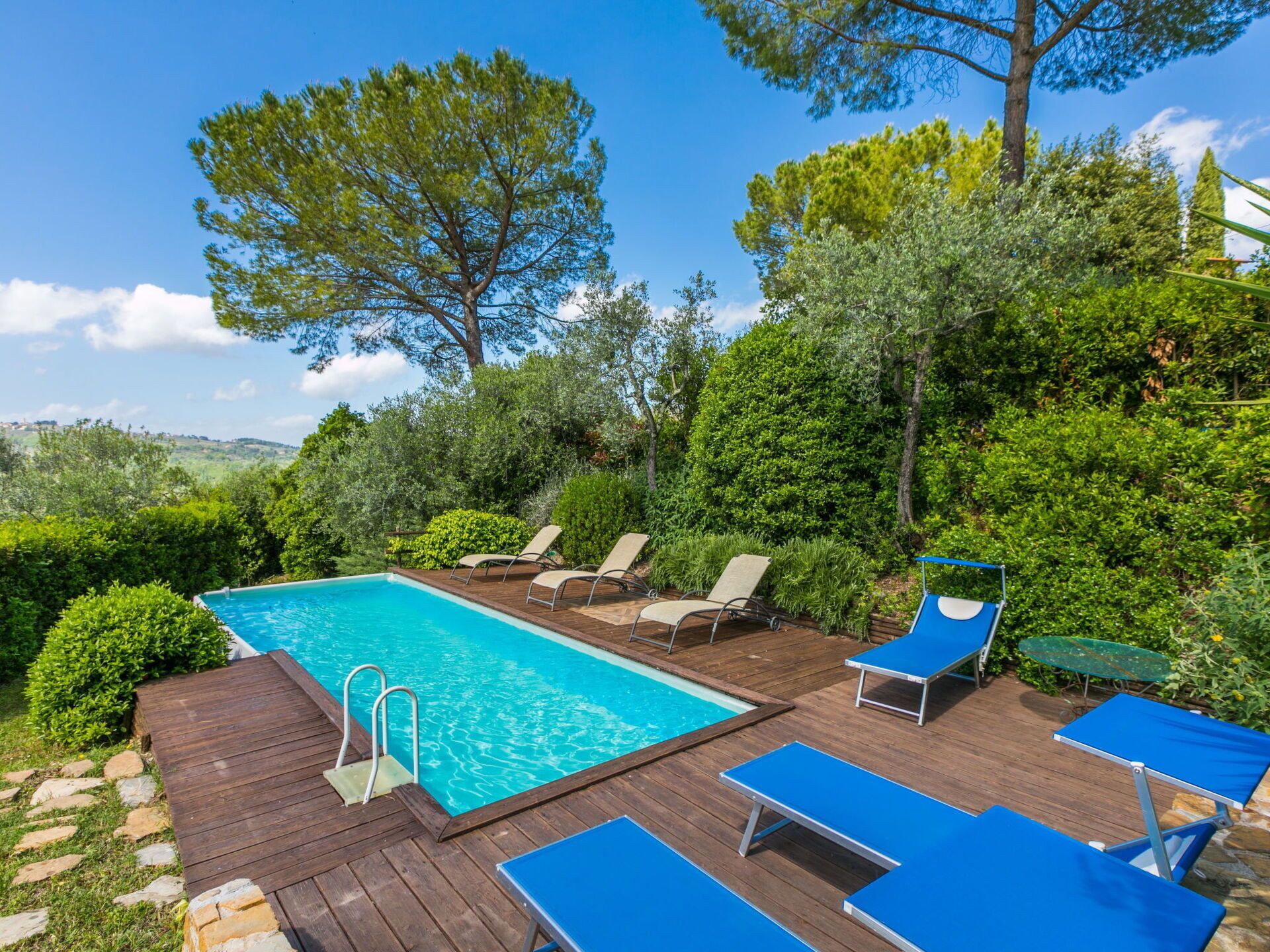 Um deck de madeira com piscina, cadeiras de descanso e vista para o verde e colinas ao redor.