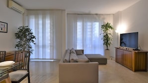 Een woonkamer met een bank, een eettafel met stoelen, een televisie op een dressoir en grote ramen met dunne gordijnen.