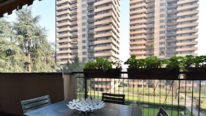 Een balkon met een tafel en stoelen, plantenbakken en uitzicht op hoge gebouwen.