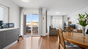 Um apartamento moderno com cozinha, sala de jantar e sala de estar. Possui uma porta de vidro deslizante que dá para uma varanda, uma mesa de jantar com cadeiras, um sofá e uma televisão.