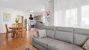 Uma sala moderna com um sofá cinzento, mesa de jantar de madeira e cadeiras. Há uma cozinha com frigorífico, forno e micro-ondas. O espaço possui paredes brancas e uma grande janela com cortinas leves.