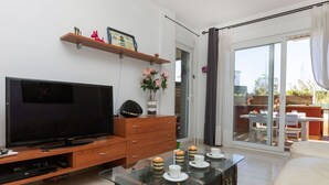 A modern living room with a flat-screen TV, wooden furniture, a glass coffee table, and a sliding glass door leading to an outdoor patio.