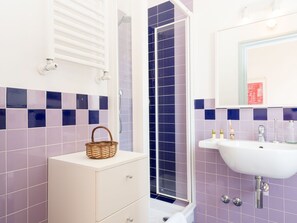 Een badkamer met een witte wastafel, een rieten mand, een douche met blauwe tegels en een wastafel met een spiegel erboven.