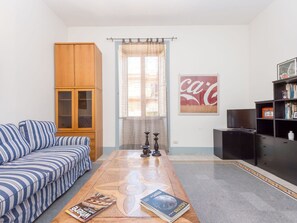 Een woonkamer met een gestreept bankje, een houten kast, een bijzettafel met tijdschriften, een boekenkast en een raam met gordijn.