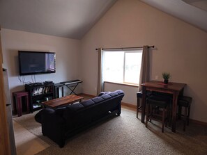 Un salon avec un canapé noir, une petite table en bois, un téléviseur fixé au mur et une fenêtre avec des rideaux.