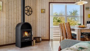 A cozy room with a wood-burning stove, wooden walls, a dining area with a table and chairs, and a large window offering a view of trees. - Beautiful home in Tarm with WiFi (Tarm)